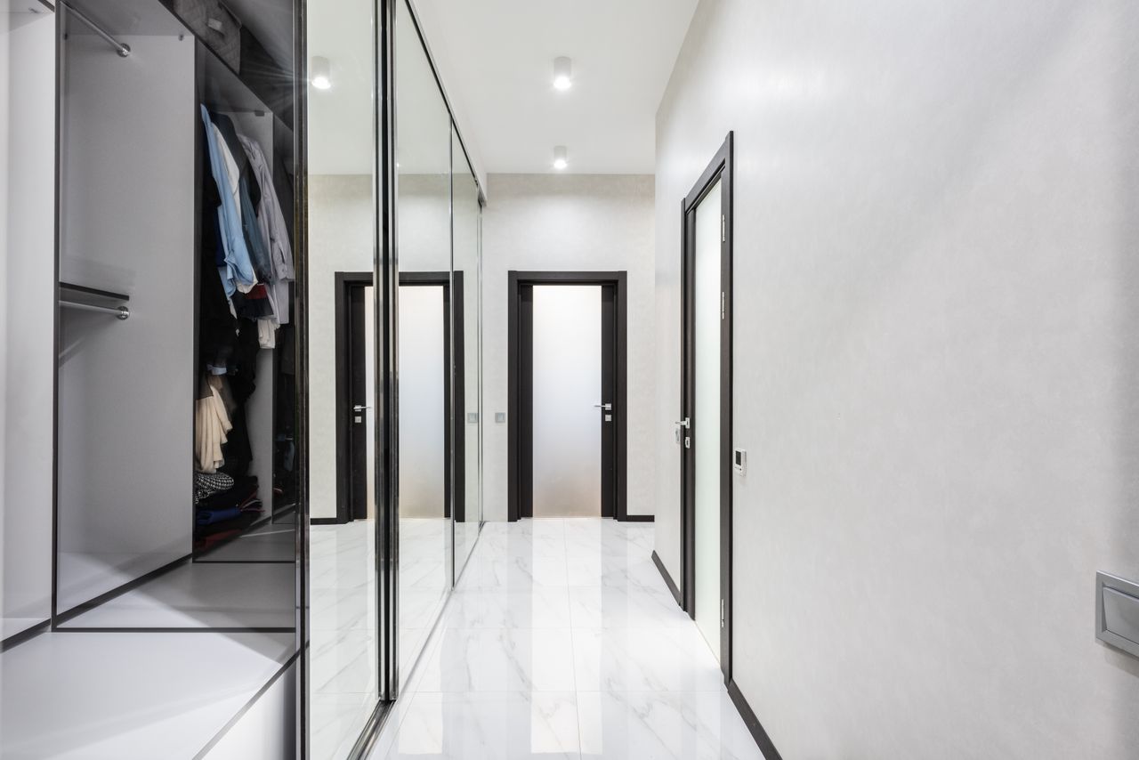 Long light corridor with white walls and opened wardrobe with cloth in modern apartment with closed doors and glowing lamps